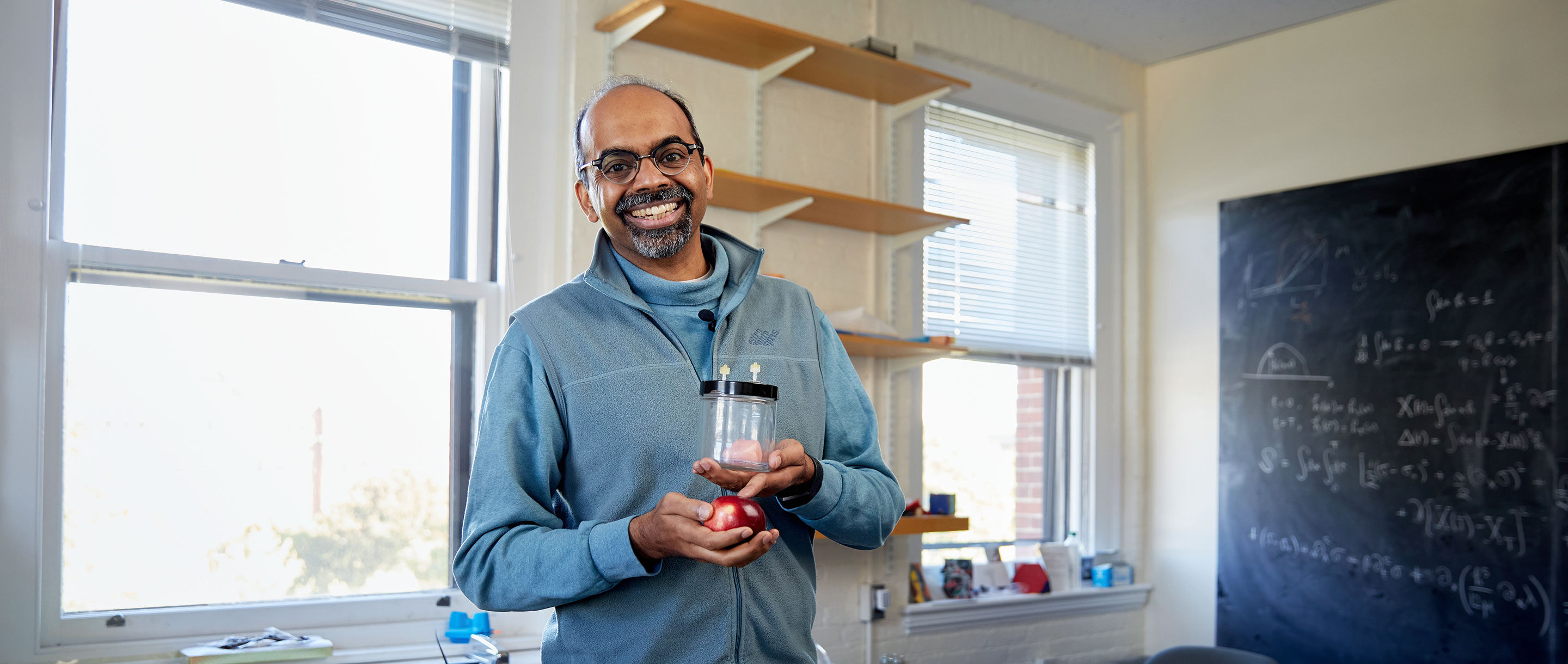 Harvard professor L. Mahadevan stands in a slightly cluttered office, holding an apple and a jar containing a miniature brain